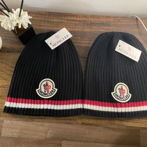 Svart och grå  ribbad mössa - Svart ribbstickad mössa  med klassisk logopatch framtill. Mössan har röd och vit rand längst ner som ger en snygg kontrast. Perfekt för dig som vill ha en stilren och exklusiv look under kalla dagar. 1 för 200 2 för 350