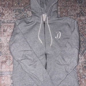 Grå hoodie med dragkedja och huva - Grå hoodie från Dobby med vit snörning i huvan och en broderad vit bokstav 'D' på bröstet. Tröjan har dragkedja framtill, långa ärmar och ribbade muddar. Perfekt för chill dagar och enkel att matcha med jeans eller joggers.