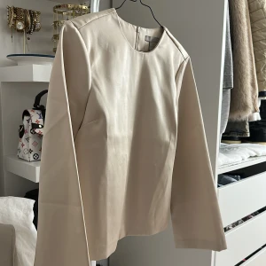 Champagnefärgad satinblus från H&M - Säljer en stilren champagnefärgad blus från H&M i satinliknande material. Blusen har rund halsringning, långa ärmar och en diskret dragkedja i nacken. Perfekt för dig som gillar en clean och elegant look.