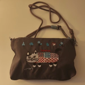 Brun axelväska med broderad katt - Säljer en brun axelväska från Jiefelai med broderad katt och färgglada detaljer på framsidan. Väskan har justerbar axelrem i repstil och dragkedja upptill. Materialet är mjukt och syntetiskt, perfekt för dig som gillar unika accessoarer med lekfull design.