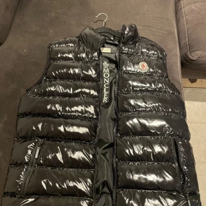 Svart dunväst från Moncler, storlek L - Snygg svart dunväst från Moncler i glansigt material med quiltad design. Västen har hög krage, dragkedja framtill och två fickor med dragkedja. Klassisk Moncler-logga på bröstet och broderad logotyp på insidan. Perfekt för lager-på-lager-stil.