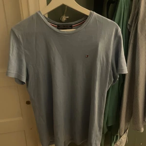 Ljusblå t-shirt från Tommy Hilfiger - Snygg ljusblå t-shirt från Tommy Hilfiger med klassisk logga broderad på bröstet. T-shirten har rund halsringning och korta ärmar. Tillverkad i mjuk bomull för en skön känsla. Perfekt basic-plagg med stilren design.