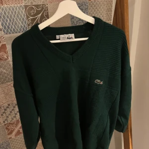 Mörkgrön stickad Lacoste tröja L - Mörkgrön stickad tröja från Lacoste med klassisk v-ringning och broderad krokodil-logga på bröstet. Tröjan har ribbade detaljer vid hals och axlar samt långärmad passform. Perfekt för dig som gillar retro och sportig stil.