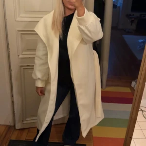 Vit lång kappa med bälte - Säljer en stilren vit kappa med bred krage och bälte i midjan. Jackan har långa ärmar med puffdetalj vid mudden och är helvit utan mönster. ALDRIG använd!