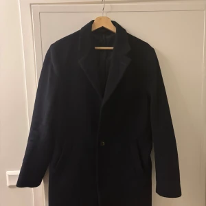 Mörkblå ullrock från John Henric - Snygg mörkblå rock från John Henric med klassisk krage och knappstängning. Tillverkad i finaste ull från Italien, med slät insida och diskreta fickor. Perfekt för dig som vill ha en stilren och tidlös look under höst och vinter. Den är mer eller mindre oanvänd. Hör av er vid frågor! Nypris är 4000kr