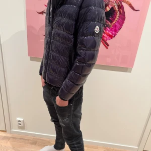 Mörkblå Moncler pufferjacka herr - Säljer en snygg mörkblå pufferjacka från Moncler med glansig finish och klassisk logga på ärmen. Jackan har quiltad design, dragkedja framtill och långärmade armar. Perfekt för dig som vill ha en stilren och varm jacka med streetkänsla.
