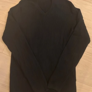 Svart ribbad stickad tröja v-ringad - Svart stickad tröja i medium med ribbad struktur och v-ringad hals. Långärmad och mjuk, perfekt för dig som gillar enkel och stilren look. Passar dig som vill ha en klassisk svart tröja med lite extra detaljer i stickningen.