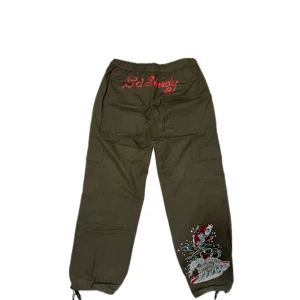 Ed Hardy x Urban Outfitters cargopants - Mörkgröna cargopants från Ed Hardy x Urban Outfitters med broderad fisk och vågor på benet, röda tryckdetaljer och snörning i midja och benslut. Byxorna har flera fickor och en loose passform, perfekta för en streetwear-look. Använd a ca. 2 gånger.