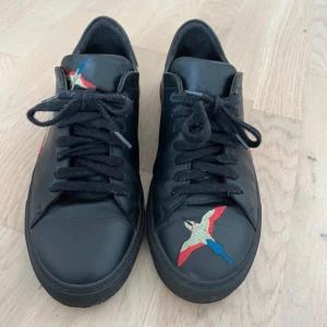 Svarta Axel Arigato sneakers med broderad fågel - Säljer mina svarta Axel Arigato-skor eftersom de tyvärr är för små för mig. Storlek 41. Skorna är i mycket bra skick och har inga defekter. Snygg design med broderade fåglar på sidorna och framtill.  Frågor eller bud är varmt välkomna – hör gärna av dig om du är intresserad! 