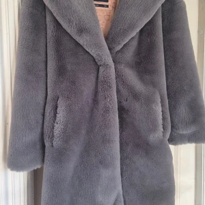 Grå fuskpälsjacka med bred krage - Mysig och fluffig grå fuskpälsjacka med bred krage och två fickor framtill. Jackan har ett lent foder med mönster på insidan och är perfekt för kyliga dagar. Långärmad och rak passform som ger en cool och trendig look. Innerfoder lite skadat. Knappar saknas men det är inget som hindrar funktionen. Stor i storleken. 