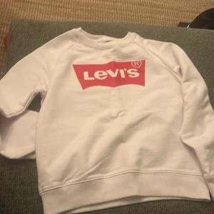 Vit sweatshirt från Levi's med röd logga - Säljer en vit sweatshirt från Levi's med den klassiska röda loggan tryckt på bröstet. Tröjan har rund hals, långa ärmar och ribbade muddar vid ärmslut och nederkant. Perfekt för dig som gillar streetwear och snygga, enkla prints.