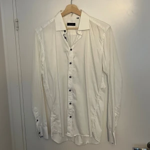 Vit skjorta från Sand Copenhagen - Vit skjorta från Sand Copenhagen i storlek 41. Klassisk modell med krage och mörka knappar framtill. Tillverkad i mjuk bomull, långärmad och med snygga detaljer vid knappslån. Perfekt för dig som gillar stilrena och enkla plagg.