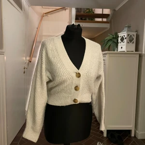 Vit croppad kofta - Supermysig vit stickad kofta med croppad passform och stora knappar framtill. Koftan har långärmade ärmar och ett ribbat mönster. Perfekt att slänga över en t-shirt eller linne när det är lite kyligt ute.