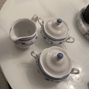 Porslinsset med blå blommor – gräddkanna & 2 sockerskålar - Charmigt porslinsset med klassiskt blått blommönster, bestående av en gräddkanna och två sockerskålar med lock. Setet är i mycket gott skick – inga synliga skador, sprickor eller nagg. Perfekt både till servering vid fika och som vacker inredningsdetalj i kök eller vitrinskåp.