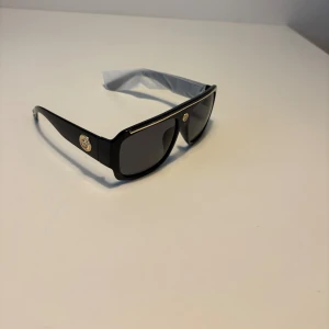 Versace liknande solglasögon  - Snygga svarta solglasögon från Versace med breda bågar och rektangulär form. De har mörka linser och guldiga Medusa-emblem på sidorna samt en tunn guldfärgad detalj längs ovansidan. Glasögonen är tillverkade i blank syntet och har en riktigt lyxig vibe. Helt nya.