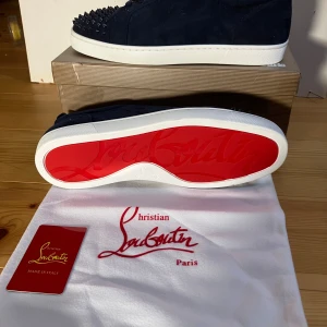 Christian Louboutin sneakers med nitar - Mörkblå sneakers i mocka från Christian Louboutin med ikonisk röd sula och vita kanter. Skorna har snörning och coola svarta nitar på tån för en edgy vibe. Perfekt för dig som vill sticka ut med lyxig streetstyle.