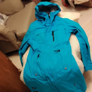 Turkos vindjacka med huva och patchar - Turkos vindjacka från Everest med huva, flera dragkedjeförsedda fickor och coola patchar på nedre delen. Jackan har justerbara ärmslut och snygga detaljer som ger en sportig vibe. Perfekt för dig som gillar färg och funktion.