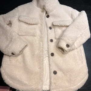 Vit teddyjacka från Lindex Kids - Mysig teddyjacka i vit färg från Lindex Kids, med stora bröstfickor och snygga knappar framtill. Jackan har lång ärm och är fodrad med slätt tyg på insidan. En liten diffus fläck framtill och på baksidan