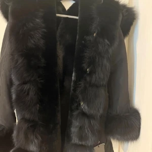 Svart pälsjacka äkta päls - Säljer en svart pälsjacka  med fluffiga pälsdetaljer längs krage, ärmslut och framkant. Jackan har en snygg och lyxig look med markerade pälsinslag som ger extra edge. Perfekt för dig som vill sticka ut med en cool och trendig jacka. Aldrig använd prislappen är kvar och är storlek S/XS pälsen är äkta.