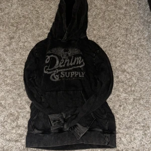 Svart hoodie från Denim & Supply Ralph Lauren - Cool svart hoodie från Denim & Supply Ralph Lauren med tryck framtill och stor huva. Tröjan har känguruficka och långa ärmar, perfekt till vilken outfit som helst. Den har en snygg vintage stil som är väldigt trendigt just nu.