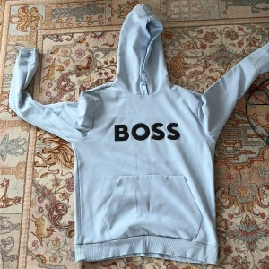 Hugo boss - Stilren och bekväm hoodie från Hugo Boss i ljusgrå färg med ikonisk BOSS-logga på bröstet. Perfekt för dig som vill ha en avslappnad men samtidigt exklusiv look. Tillverkad i mjukt och högkvalitativt material som känns skönt mot huden.