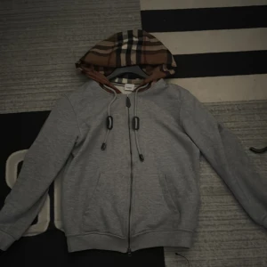 Grå hoodie från burberry - Grå hoodie med dragkedja och klassisk Burberry-inspirerad rutig huva i brunt, svart och beige. Tröjan har snörning i huvan, två fickor framtill och ribbade muddar. Perfekt för dig som gillar streetstyle och vill sticka ut med snygga detaljer.