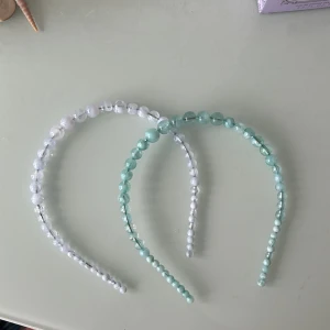 Två pärlbeströdda diadem i pastell - Säljer två snygga diadem med stora pärlor i plast, ett i ljus mintgrön och ett i skimrande vitt. Perfekt för att ge din look en extra touch med glans och färg. Diademen har rundad form och pärlorna är genomskinliga med marmorerad effekt.