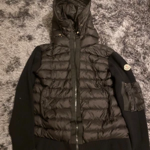 Svart cardigan från Moncler - Svart cardigan från Moncler med stickade ärmar och huva. Jackan har dragkedja framtill, snörning vid huvan och en Moncler-logga på vänster ärm. Fickor med dragkedja på sidorna och en extra ficka på ärmen. Perfekt för dig som vill ha en snygg och varm cardigan