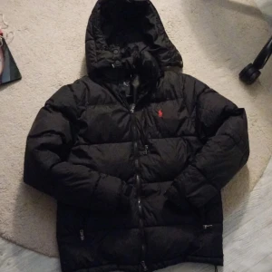 Svart dunjacka från Polo Ralph Lauren - Säljer en svart dunjacka från Polo Ralph Lauren med huva och dragkedja. Jackan har en klassisk pufferstil och är försedd med Polo-loggan broderad i rött på bröstet. Perfekt för kalla dagar och riktigt snygg till streetwear-looken.