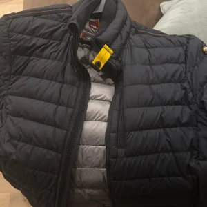 Svart dunjacka från Parajumpers - Säljer en svart dunjacka från Parajumpers med klassisk gul detalj vid kragen och snyggt broderat märke på ärmen. Jackan har quiltad design, dragkedja och lång ärm. Insidan är ljusgrå och jackan är perfekt för kalla dagar.