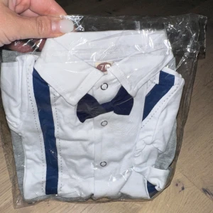 Babykostym med fluga och hängslen - Supersöt babykostym med vit överdel, blå fluga och blå hängslen. Nedre delen är grå med tryckknappar längs benen för enkel påklädning. Skjortkrage och långärmad design ger en klassisk look. Perfekt för små stunder med stil. Söt babybody med skjortdesign, komplett med fluga och hängslen. Perfekt för speciella tillfällen eller bara för att se extra stilig ut.