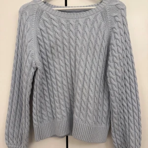 Kabelstickad tröja från Part Two - Säljer en ljusgrå stickad tröja från Part Two med grov kabelstickning och rund hals. Tröjan har långa ärmar och breda ribbade muddar vid ärmslut och nederkant. Perfekt för dig som gillar en chill och cozy stil under kyliga dagar.
