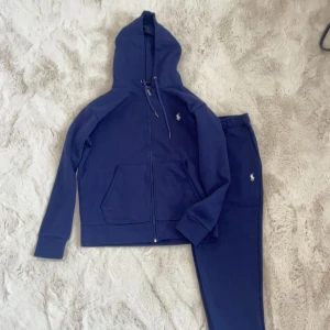Mörkblå hoodie och byxor från Polo Ralph Lauren - Säljer en snygg och bekväm mörkblå hoodie och matchande byxor från Polo Ralph Lauren. Hoodien har dragkedja och en liten vit logga på bröstet. Byxorna har en elastisk midja med dragsko och samma vita logga på benet. Perfekt för en avslappnad dag eller träning. Båda plaggen är i nyskick och gjorda av mjuk bomull. 
