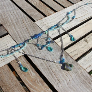 Vackert halsband med blå och gröna pärlor - Säljer ett superfint halsband med en mix av blå och gröna pärlor. Halsbandet har flera kedjor och hängande droppformade pärlor i olika nyanser av blått och grönt. Perfekt för att ge en färgklick till din outfit!