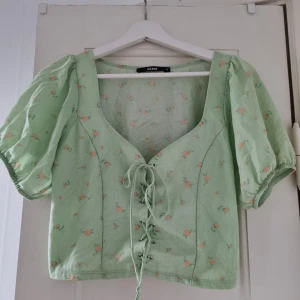 Grön blommig topp med snörning från Bik Bok - Säljer en superfin grön topp från Bik Bok med små blommor i rosa och orange. Toppen har korta puffärmar och en snygg snörning framtill. Perfekt för sommaren och vårens alla fester! 🌸 