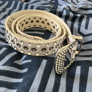Beige bälte med nitar och strass - Säljer ett snyggt beige bälte med massor av nitar och strassdetaljer. Bältet har en cool och glittrig stil som passar perfekt för festliga tillfällen. Spännet är också dekorerat med strass vilket ger en extra touch av glamour. Perfekt för att piffa upp vilken outfit som helst!
