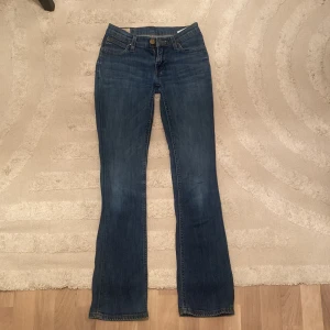 Blå jeans från Lee - Säljer ett par snygga blå jeans från Lee. De är i bra skick och har en klassisk femficksdesign med en knapp och dragkedja framtill. Jeansen har en bootcut-stil och är perfekta för både vardag och fest. De är tillverkade i ett slitstarkt denim-material som håller länge.