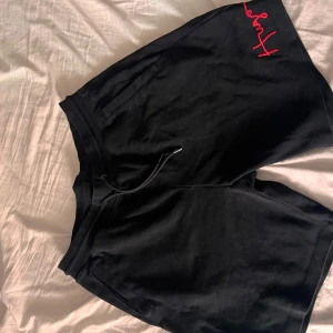 Svarta shorts från Hugo Boss - Säljer ett par svarta shorts från Hugo Boss. De är riktigt bekväma och har en snygg röd Hugo-logga på ena benet. Shortsen har dragsko i midjan och två fickor på sidorna. Perfekta för en avslappnad dag eller träning!