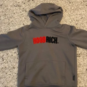 Grå hoodie från Hoodrich - Säljer en snygg grå hoodie från Hoodrich. Den har en stor ficka på magen och texten 'Hoodrich' broderad i rött och svart på bröstet. Perfekt för höst och vinter, superbekväm och stilren. Passar bra till både vardag och träning.