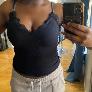 Svart ribbad linnetopp med spetsdetaljer - Säljer en svart ribbad linnetopp i storlek M. Toppen har smala axelband och fina spetsdetaljer runt halsringningen. Perfekt för sommaren eller som ett lager under en tröja. Toppen är i perfekt skick och väldigt bekväm att ha på sig.