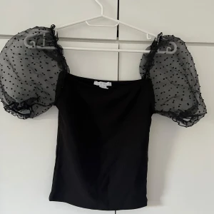 Svart topp med puffärmar - Säljer en superfin svart topp med puffärmar i genomskinligt material med små prickar. Toppen är tight och perfekt för festliga tillfällen. Den har en elegant och modern stil som verkligen sticker ut. Passar perfekt till både kjol och byxor!