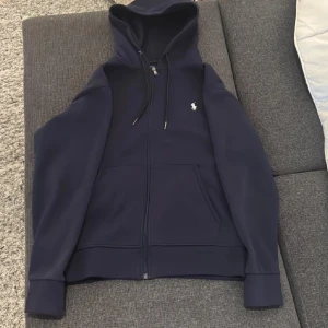 Mörkblå hoodie från Polo Ralph Lauren - Säljer en snygg och bekväm mörkblå hoodie från Polo Ralph Lauren. Den har en dragkedja framtill, en praktisk ficka och en liten vit logga på bröstet. Perfekt för höst och vinter! 🧥