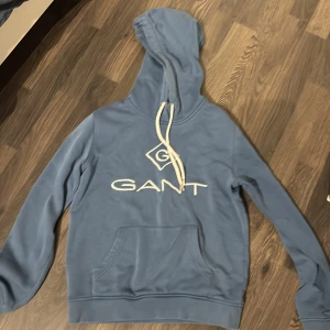 Blå hoodie från GANT - Säljer en snygg blå hoodie från GANT med vit logga på bröstet. Tröjan har en klassisk känguruficka och justerbar huva med vita snören. Perfekt för en avslappnad stil.