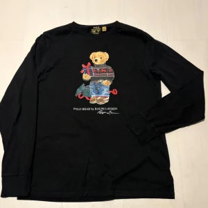 Svart långärmad tröja från Ralph Lauren - Säljer denna långärmade tröjan från Ralph lauren med en stilren Teddy bear på magen, jag säljer denna för att den inte passar längre. Den är storlek S och skulle säga att den passar en som är 170. Hör av er funderingar och pris kan diskuteras vid snabb affär!