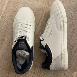 Vita sneakers från Pepe Jeans - Snygga vita sneakers från Pepe Jeans med marinblå detaljer och snörning. Skorna har en stilren design med märkets logga på sidan och en bekväm sula. Perfekta för en avslappnad stil. Passar perfekt till ett par mörka elr blåa jeans. *aldrig använda 