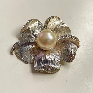 Silverfärgad blomformad brosch med pärla - Elegant brosch i form av en blomma med en stor pärla i mitten. Bladen är silverfärgade med glittrande detaljer längs kanterna. Perfekt accessoar för att ge en touch av glamour.
