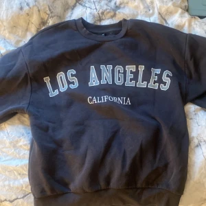 Svart sweatshirt med Los Angeles-tryck - Säljer en svart sweatshirt med texten 'Los Angeles California' tryckt på framsidan. Tröjan har långa ärmar och en rund halsringning. Säljs på grund av för liten storlek 