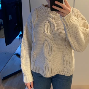 Vit stickad tröja med flätmönster - Naturvit stickad tröja med ett flätmönster. Tröjan har långa ärmar och en ribbad krage. 