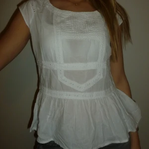 Vit blus med spetsdetaljer - Säljer en söt vit blus med spetsdetaljer och korta ärmar. Blusen har en fin passform och en volangkant nedtill. Perfekt för en uppklädd look!