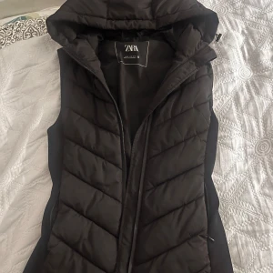 Svart dunväst från Zara - Snygg svart dunväst från Zara med huva och dragkedja. Perfekt för kyliga dagar när du vill hålla stilen. Västen har en quiltad design som ger en modern look.